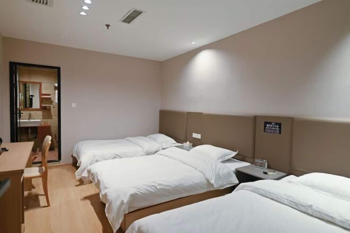 A hotel room with three single beds, a desk, a chair, and a small table with a lamp.