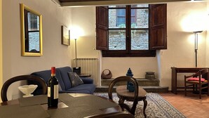 Un salon chaleureux avec un canapé, une table à manger agrémentée d’une bouteille de vin, une lampe et une vue sur un mur de pierre, visible à travers une fenêtre.