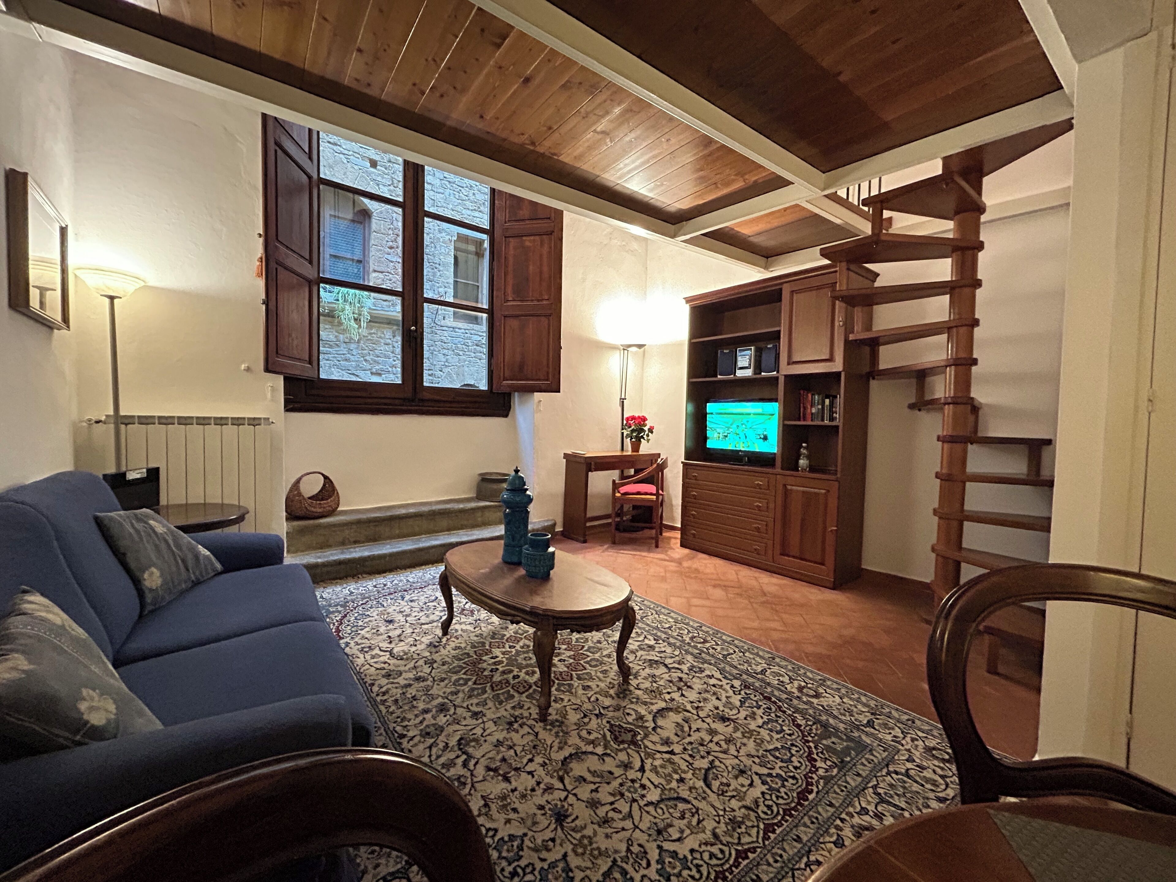 A cozy living room with a blue sofa, a wooden coffee table, a patterned rug, a television, and a wooden staircase.