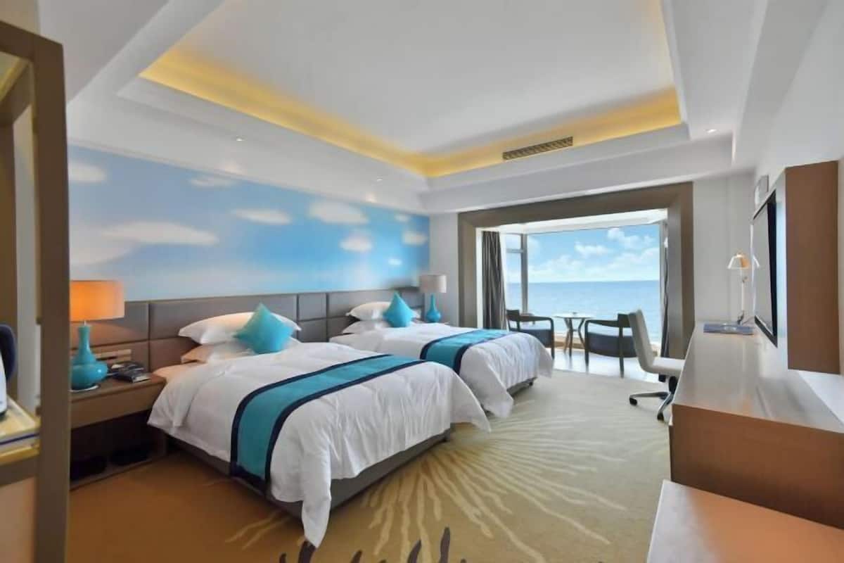 Ein Hotelzimmer mit zwei Betten, einem Schreibtisch, einem Stuhl und einem großen Fenster mit Meerblick.