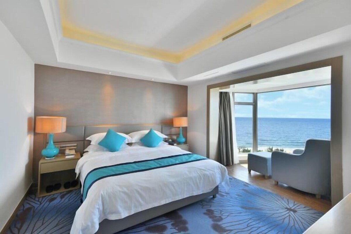 A modern hotel room with a large bed, two bedside lamps, a view of the sea, and a seating area with a view.