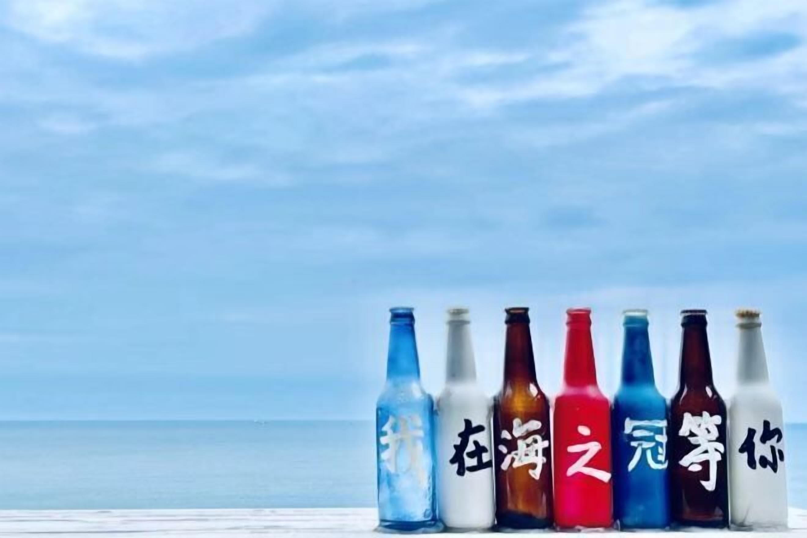 Seis botellas con caracteres chinos sobre una superficie de madera, con el mar y la playa al fondo.