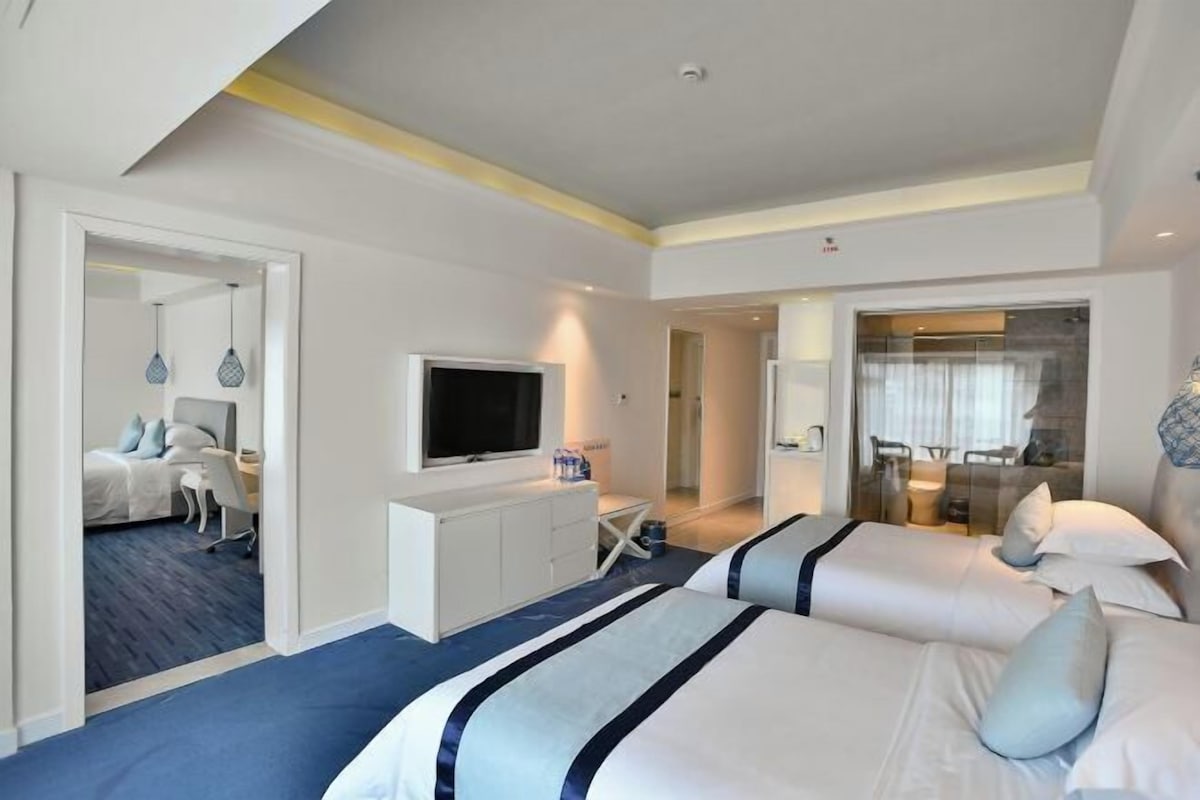 A modern hotel room with two beds, a TV, a desk, and a dining area visible through an open door.