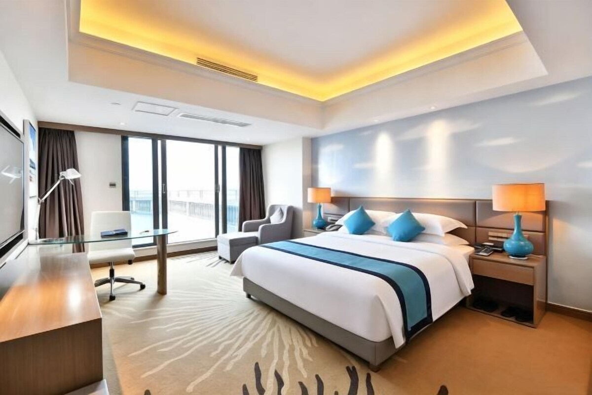 A hotel room with a large bed, a desk, a chair, a TV, and a view of the outdoors.