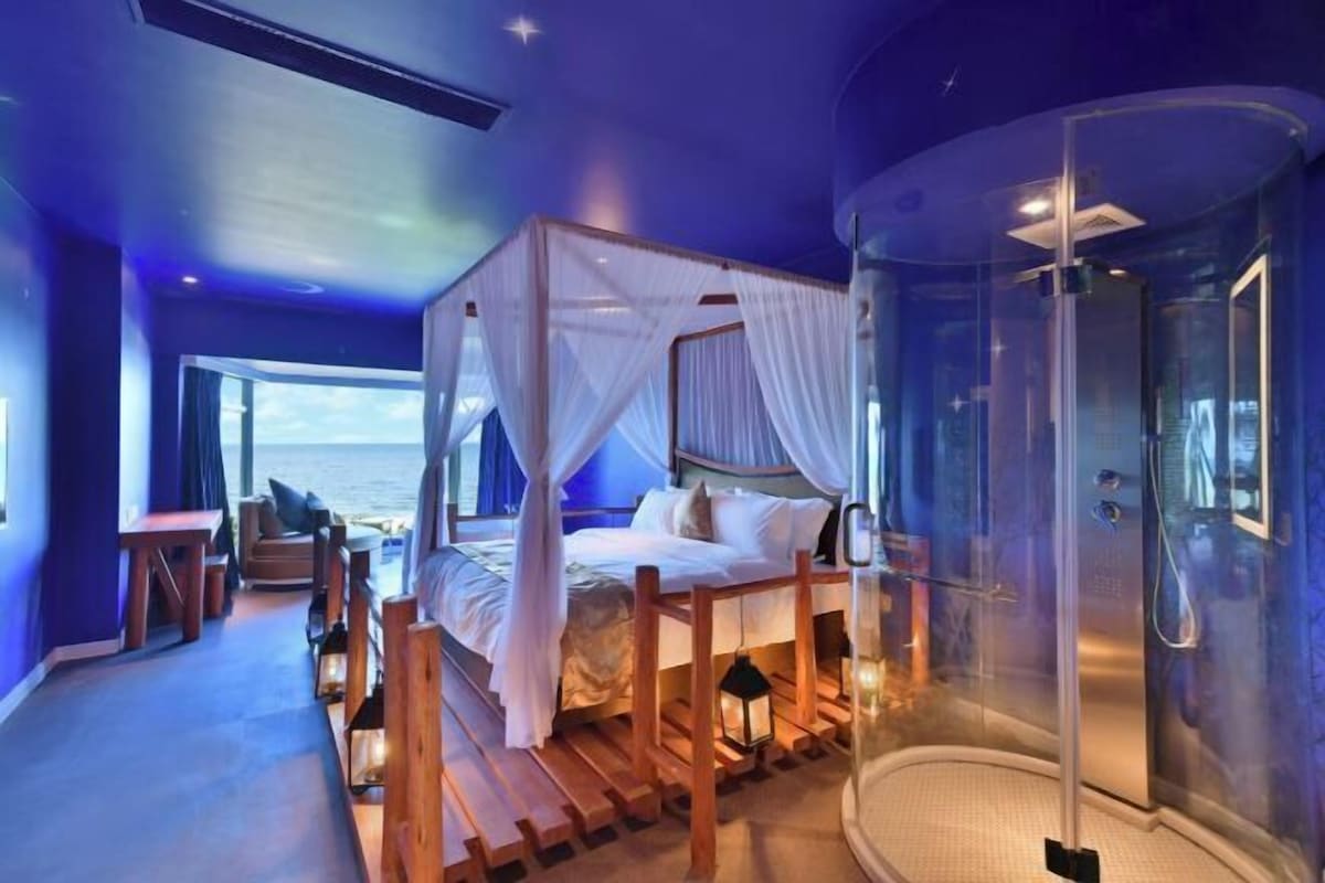 A bedroom with a four-poster bed, a glass-enclosed shower, and a view of the sea.
