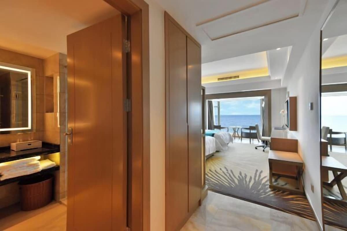 A hotel room with a large bed, a desk, a chair, a bathroom with a mirror, and a view of the sea.