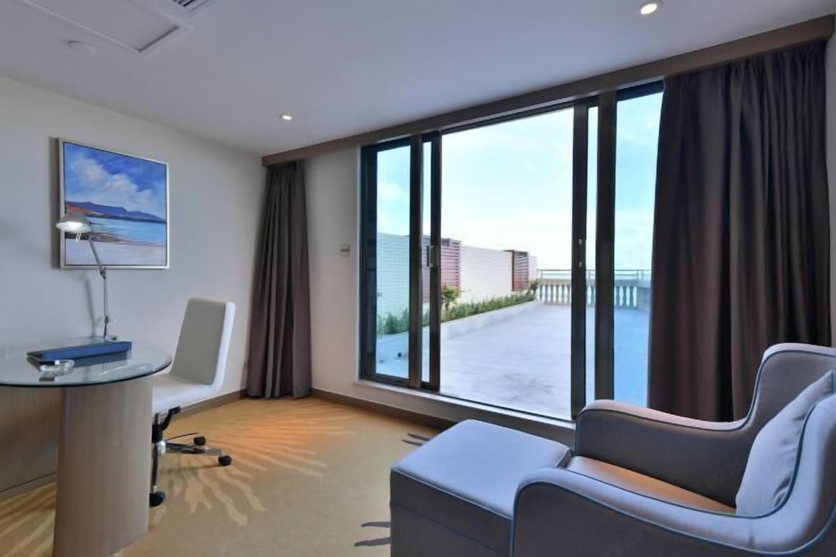 A hotel room with a large window, a desk with a lamp, a chair, and a sofa.