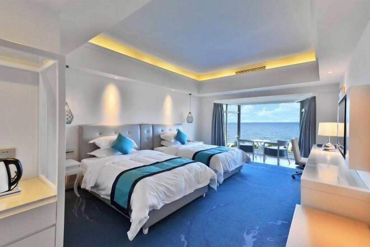 Ein Hotelzimmer mit zwei Betten, einem Schreibtisch, einem Sessel, einer Kaffeemaschine und Blick auf das Meer.