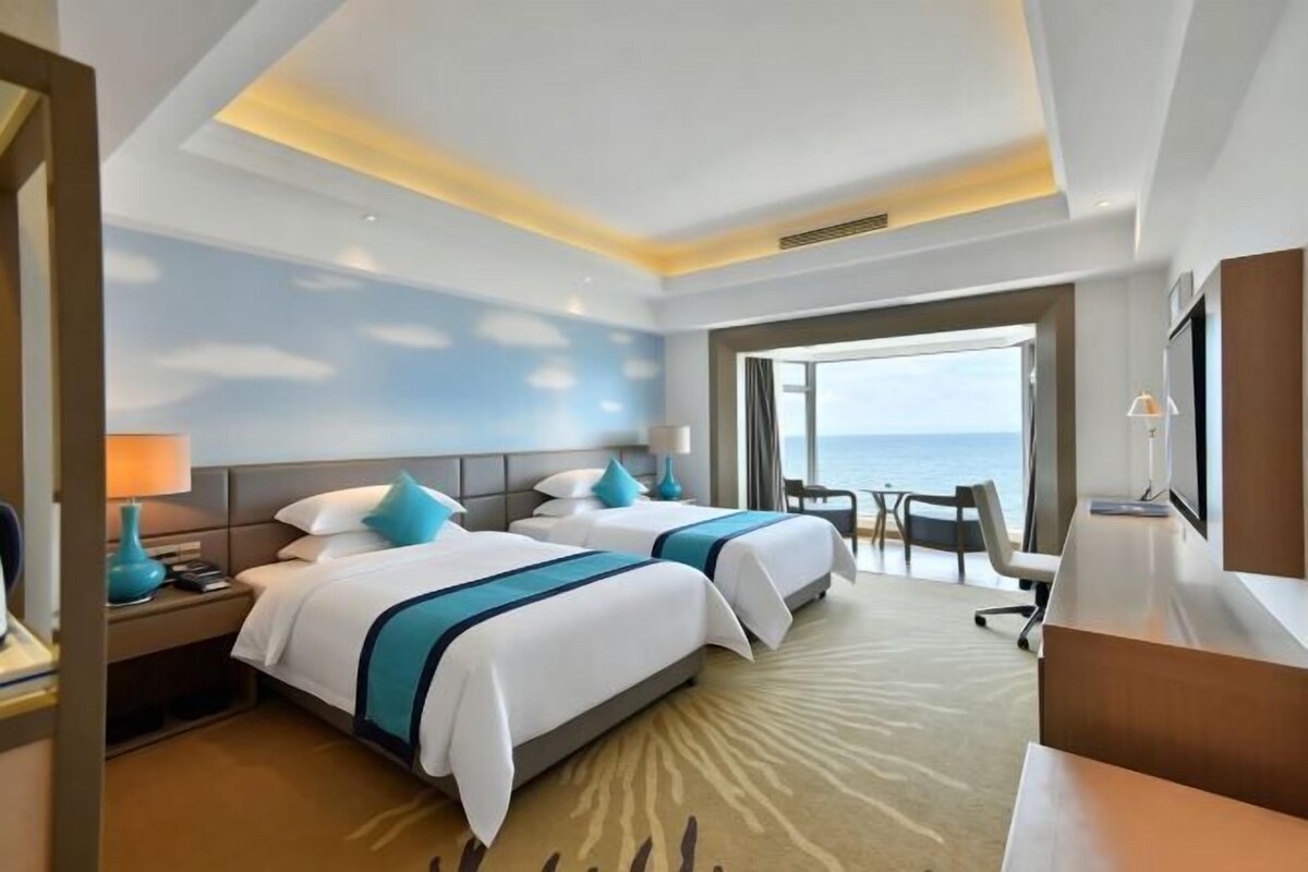 A hotel room with two beds, a desk, a chair, and a view of the sea.