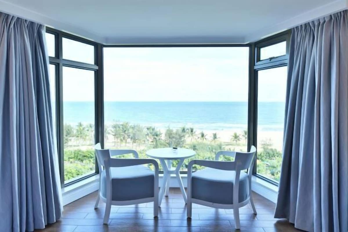 A room with a large window overlooking the sea, two chairs, and a small table.