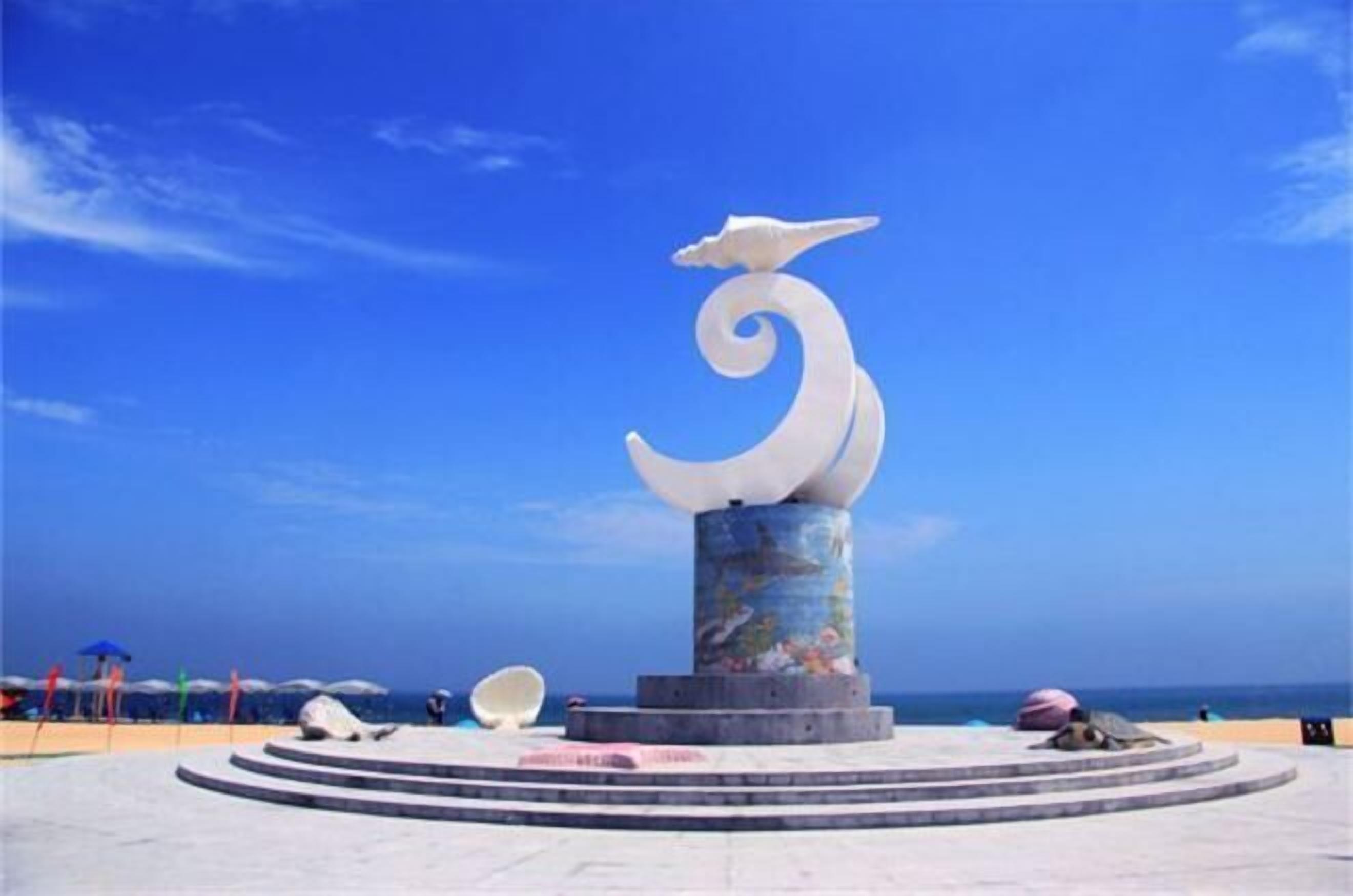 Eine Skulptur mit Vogelmotiv und spiralförmigem Design, platziert auf einer runden Plattform mit Stufen, vor einem strahlend blauen Himmel.