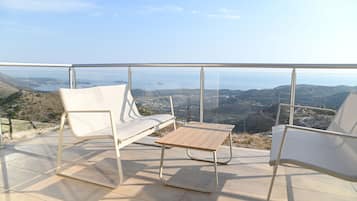 A balcony with two white chairs and a small table, overlooking a scenic landscape with hills and a body of water.