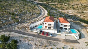 Un edificio moderno de dos pisos con piscina, una entrada curva y un estacionamiento con varios coches.