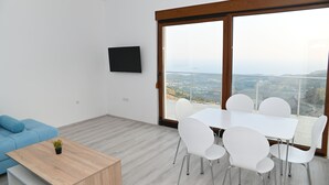 Un salon moderne doté d’une grande fenêtre, d’un téléviseur à écran plat, d’un canapé bleu, d’une table basse en bois et d’une table à manger blanche avec des chaises.