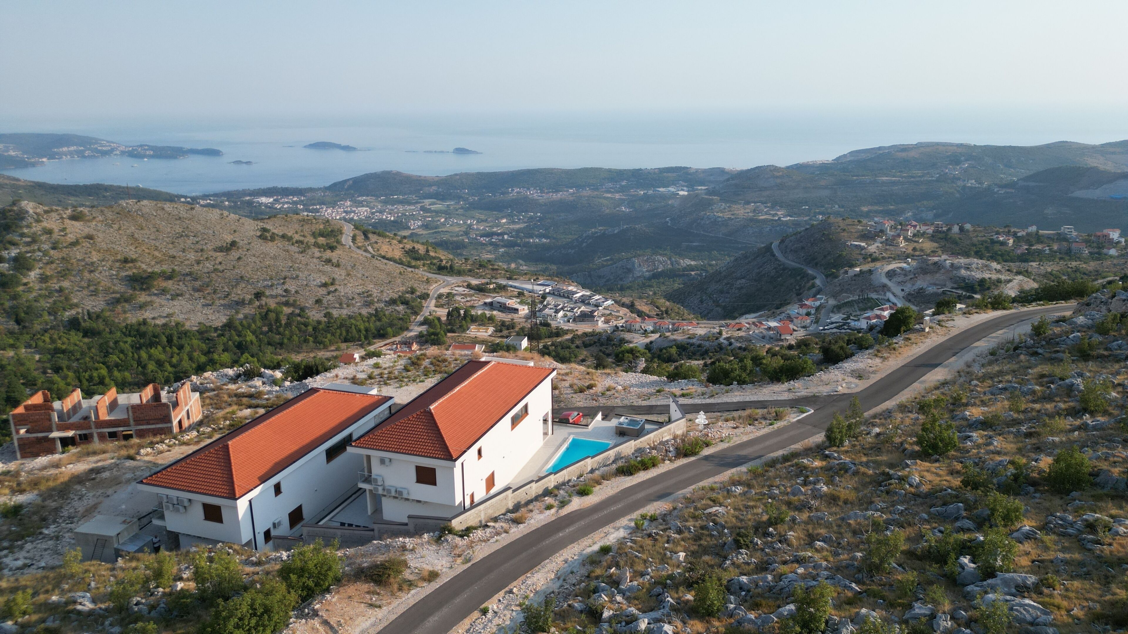 A coastal view with a road leading to a white building with a red roof, surrounded by rocky terrain and distant hills.