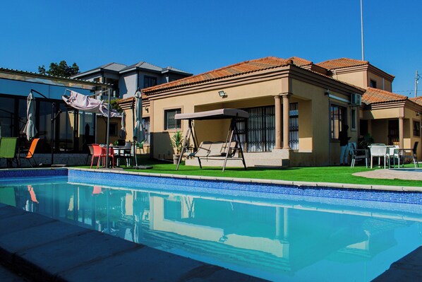 Una piscina con sillas de descanso y sombrillas, rodeada por un césped cuidado y un edificio con techo de tejas.