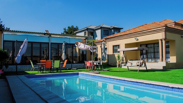 Una piscina con sillones de playa y sombrillas, rodeada de un césped cuidado y un edificio con ventanas amplias.