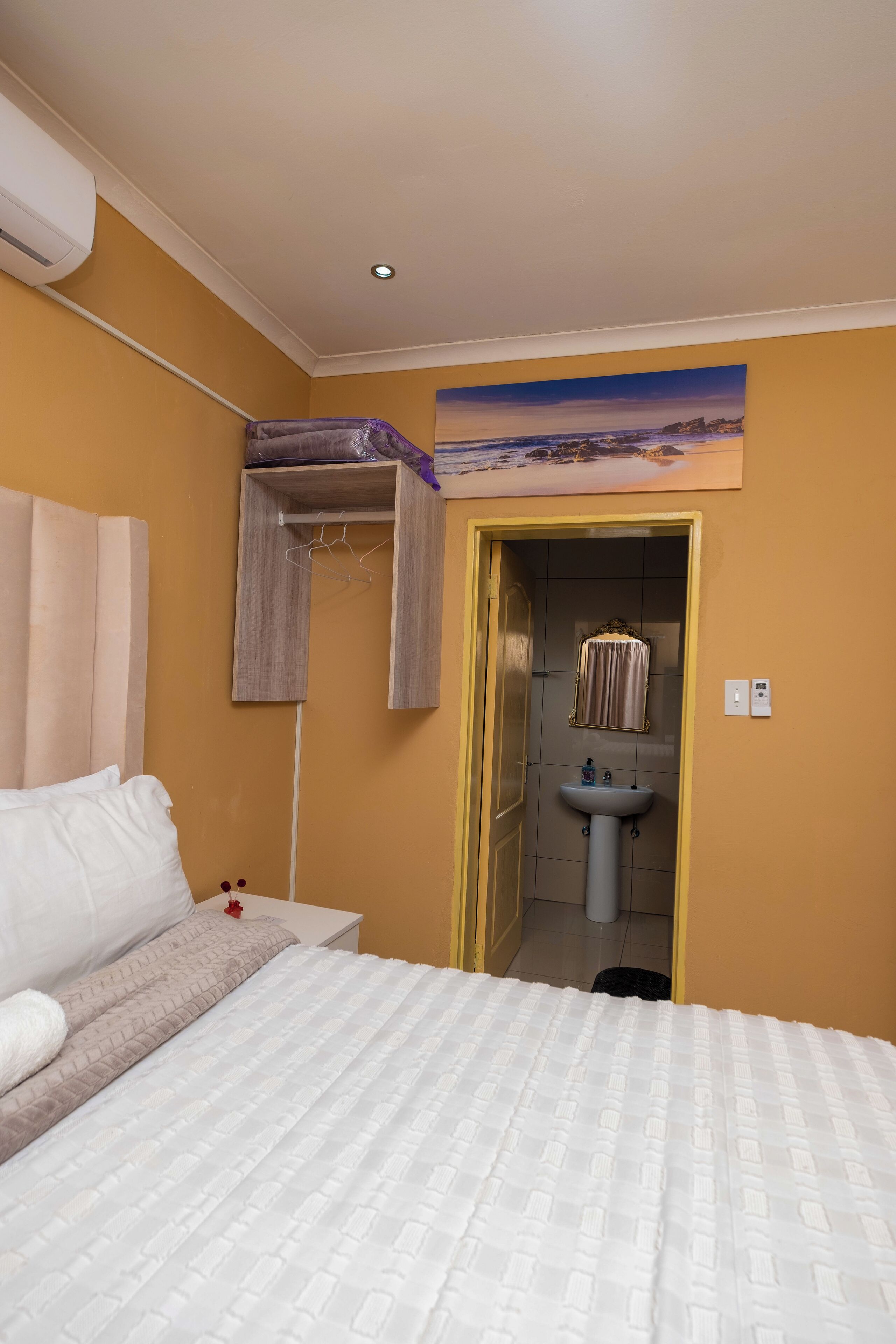 Habitación de hotel con una cama, una mesita de noche, un armario, un baño con lavamanos y un cuadro en la pared.