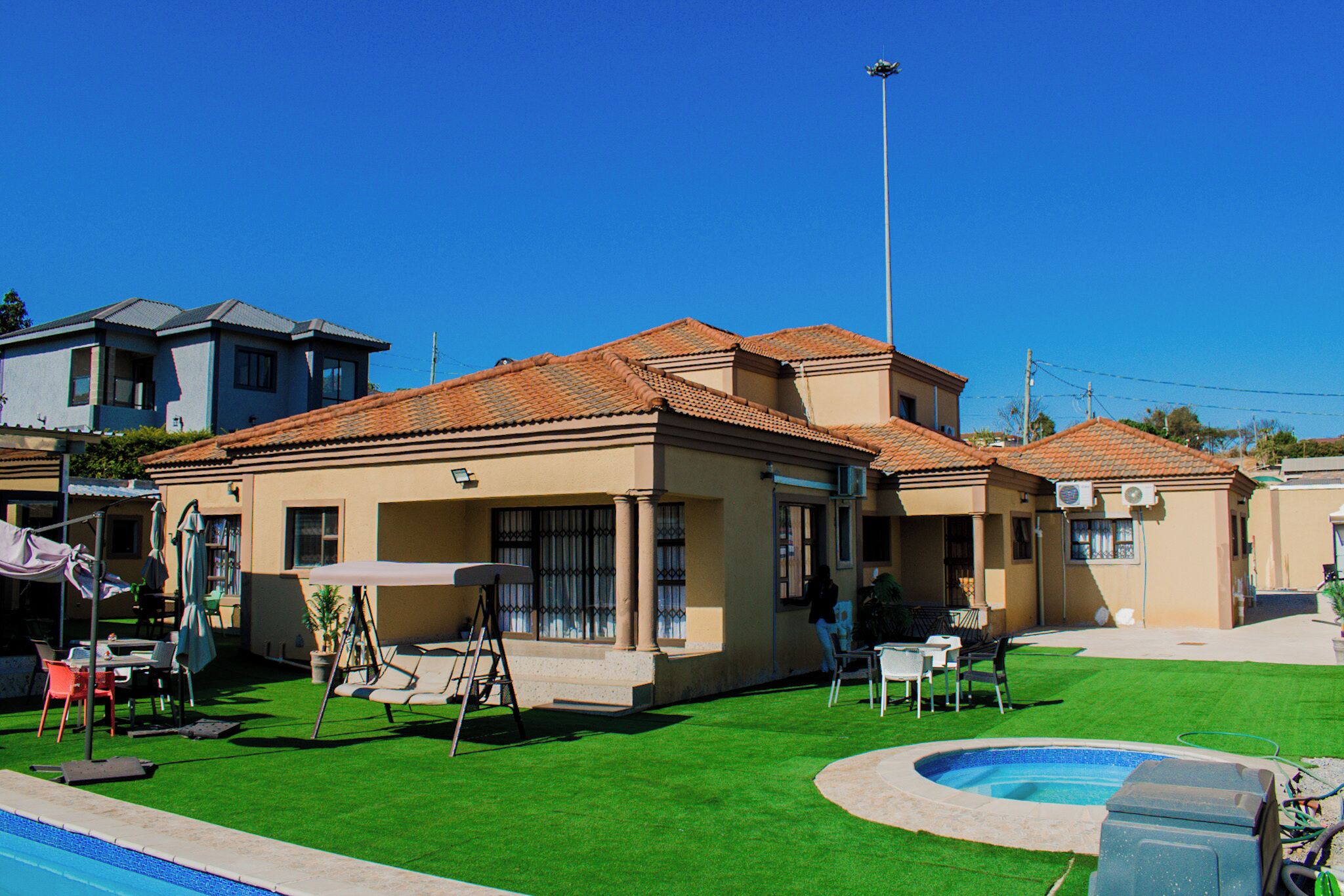 A residential area with a swimming pool, green artificial grass, and a house with a tiled roof.
