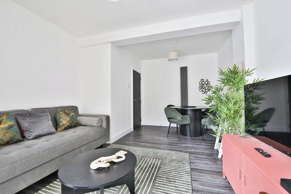 A modern living room with a grey sofa, a round coffee table, and a red cabinet. There is a dining area with a table and chairs, and a large green plant.
