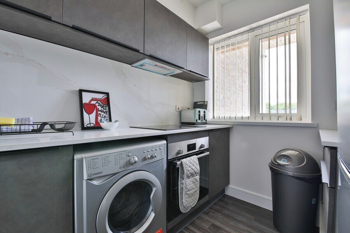 A compact kitchen with a washing machine, oven, and a window with blinds.