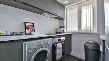 A compact kitchen with a washing machine, oven, and a window with blinds.