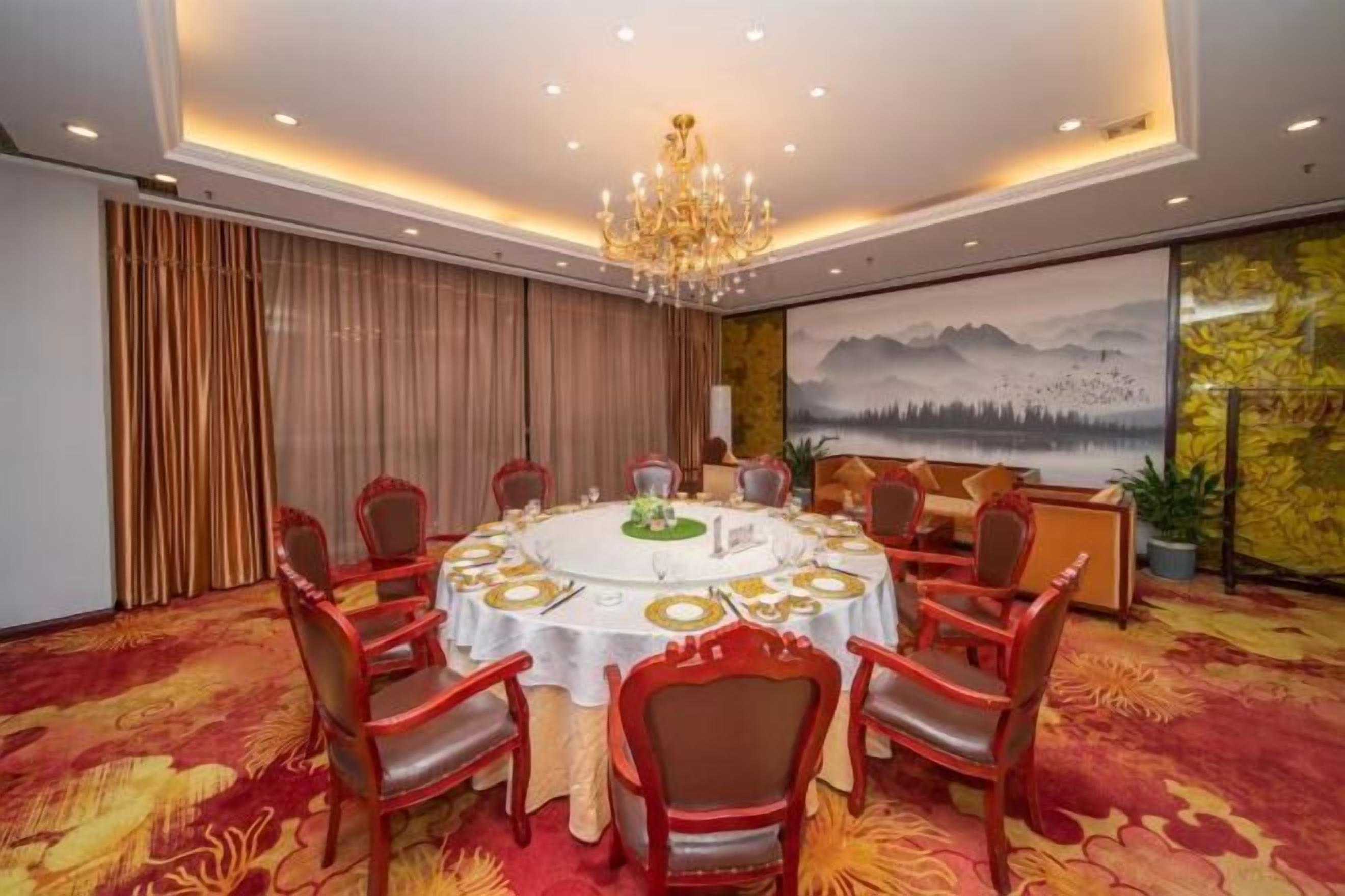 A conference room with a round table, red chairs, a chandelier, and a painting of mountains.