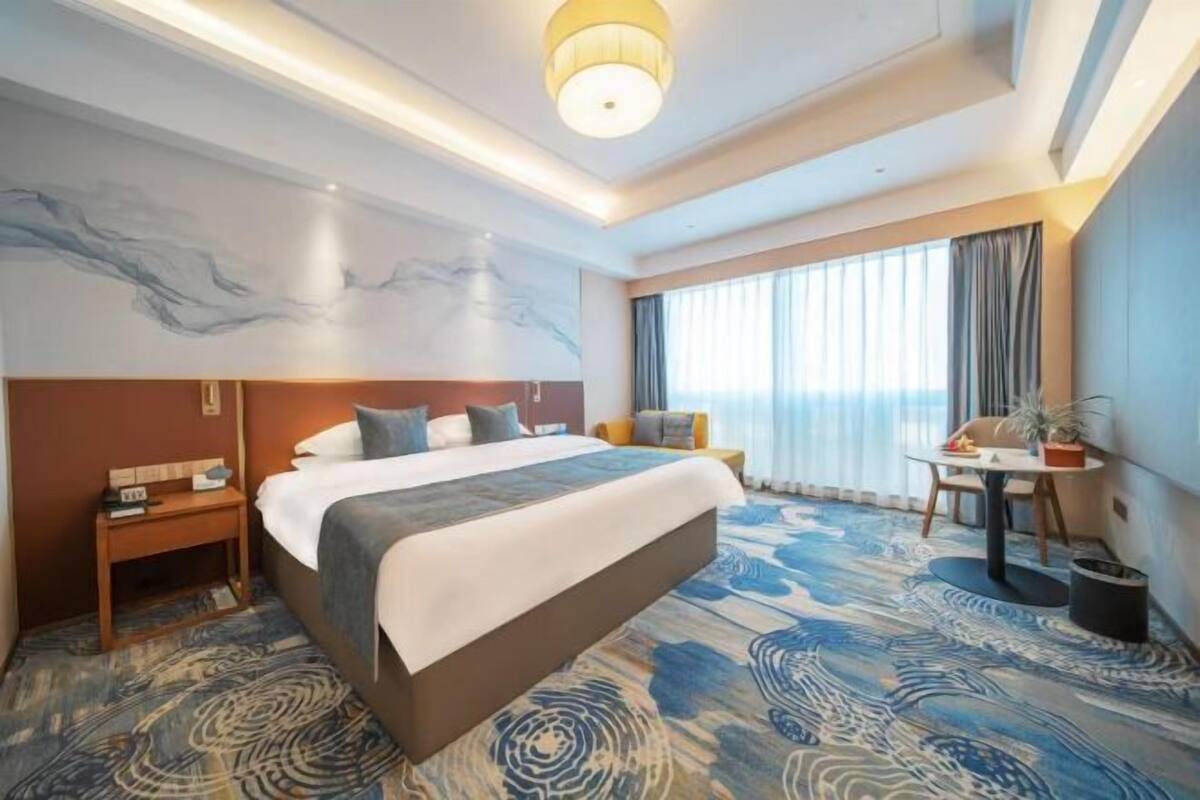 Ein Hotelzimmer mit einem großen Bett, einem Schreibtisch, einem Sessel, einem kleinen Tisch und einem großen Fenster mit Vorhängen.