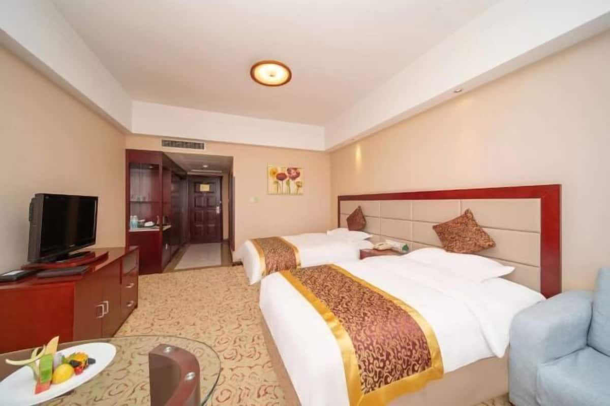 Ein Hotelzimmer mit zwei Betten, einem Fernseher, einem Schreibtisch mit Stuhl und einem Esstisch mit einer Obstplatte.