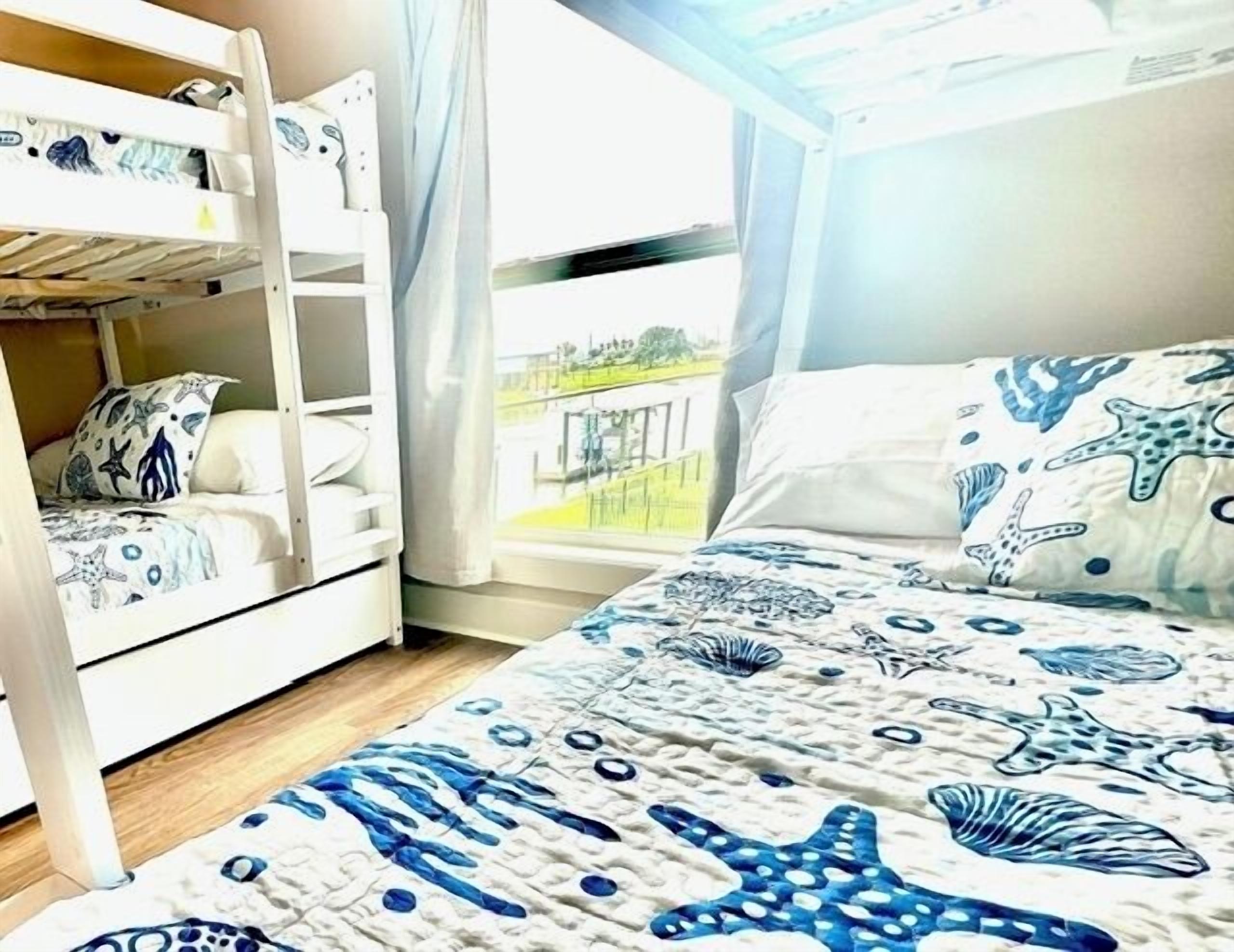 Een kamer met stapelbedden, een groot bed met sterren en schelpen motieven, en een raam met uitzicht op een hek en begroeiing.