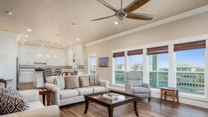A spacious living room with a sofa, armchairs, and a coffee table. A kitchen is visible in the background, and there are large windows with a view of buildings. - Boaters' paradise, direct access to the Intracoastal, East Bay and Gulf. (Bolivar Peninsula)