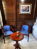 Una habitación con dos sillas de madera con cojines azules, una pequeña mesa de madera con un florero y un cuadro en la pared.