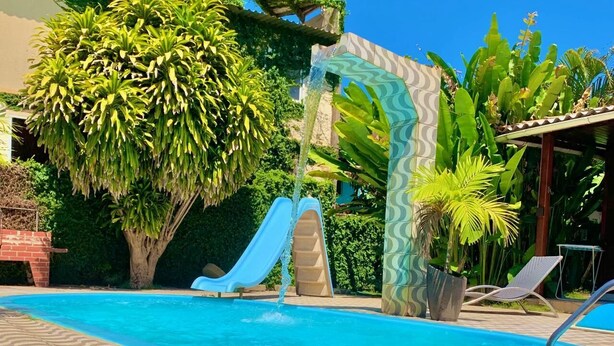 A swimming pool with a slide, surrounded by lush greenery and a tiled deck. - Pousada das Flores (Fernando de Noronha)