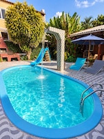 Una piscina con tobogán, rodeada de sillas de descanso y una zona de barbacoa de ladrillo.