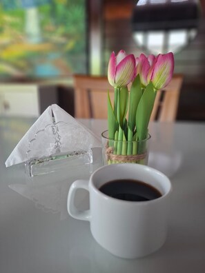 Eine transparente Glaspyramide, eine Glasvase mit rosa Tulpen und eine weiße Tasse mit Kaffee auf einem Tisch.