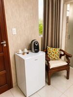 Un electrodoméstico blanco compacto, posiblemente un refrigerador pequeño, con una cafetera encima, una puerta de madera y una silla con un cojín de girasol.
