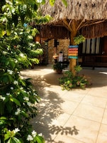 A tropical outdoor area with a thatched roof structure, potted plants, and a sign with multilingual greetings.