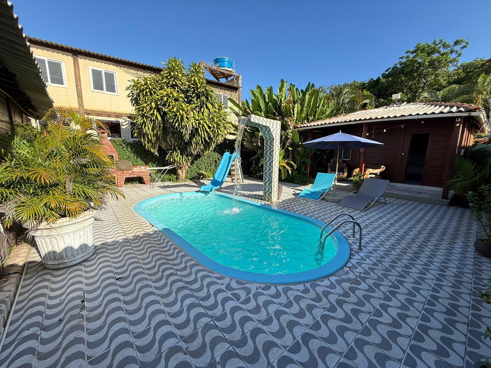 Uma piscina com escorregador, rodeada por espreguiçadeiras e um chão de azulejos estampados.