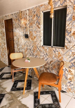 A dining area with a round table and two chairs, a window with curtains, and a wall with patterned tiles. - Pousada das Flores (Fernando de Noronha)