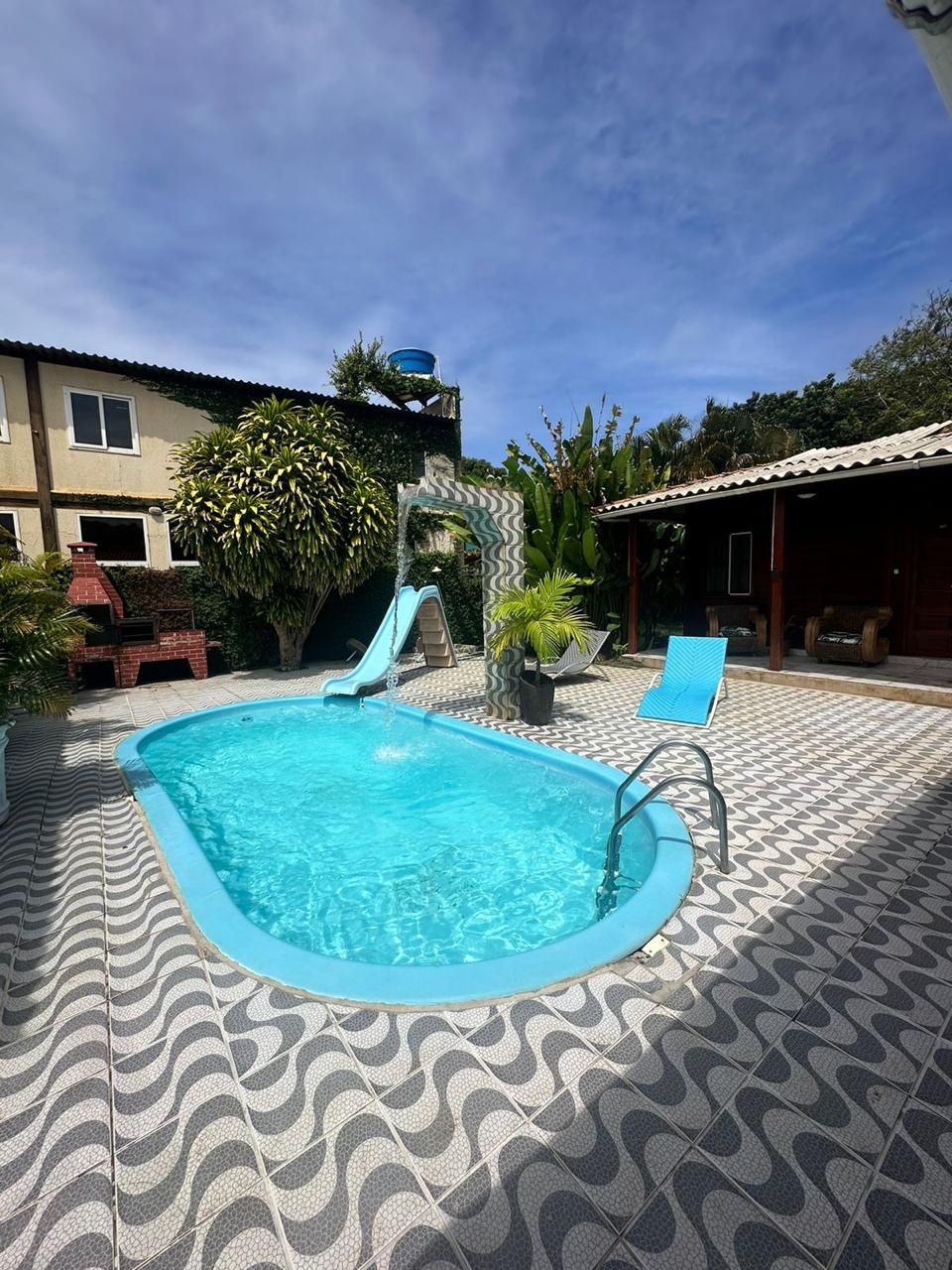 Ein Swimmingpool mit Rutsche, umgeben von einer gemusterten Terrasse und einem überdachten Bereich mit Sitzgelegenheiten.