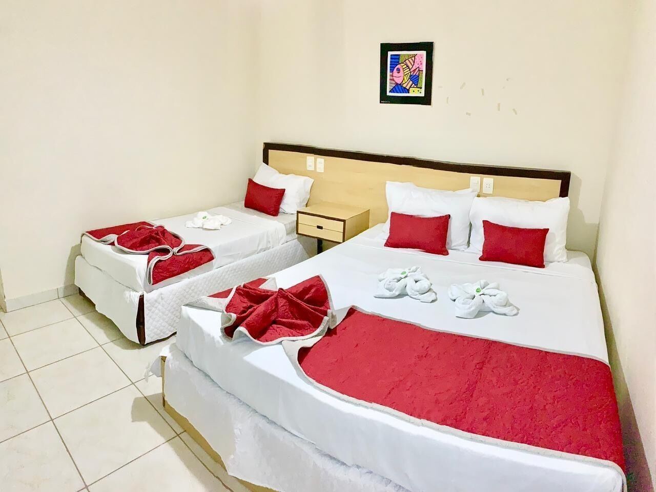 Habitación de hotel con dos camas, cada una con ropa blanca y detalles rojos, incluyendo almohadas y una colcha. Hay una mesita de noche pequeña con una lámpara y un cuadro enmarcado en la pared sobre las camas.