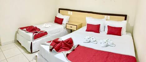 Quarto de hotel com duas camas, cada uma com roupa de cama branca e detalhes vermelhos, incluindo almofadas e um cobertor. Há uma pequena mesa de cabeceira com uma lâmpada e uma moldura com um quadro na parede acima das camas.