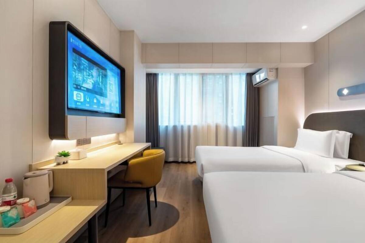 A hotel room with a desk, two beds, a TV mounted on the wall, and a window with curtains.