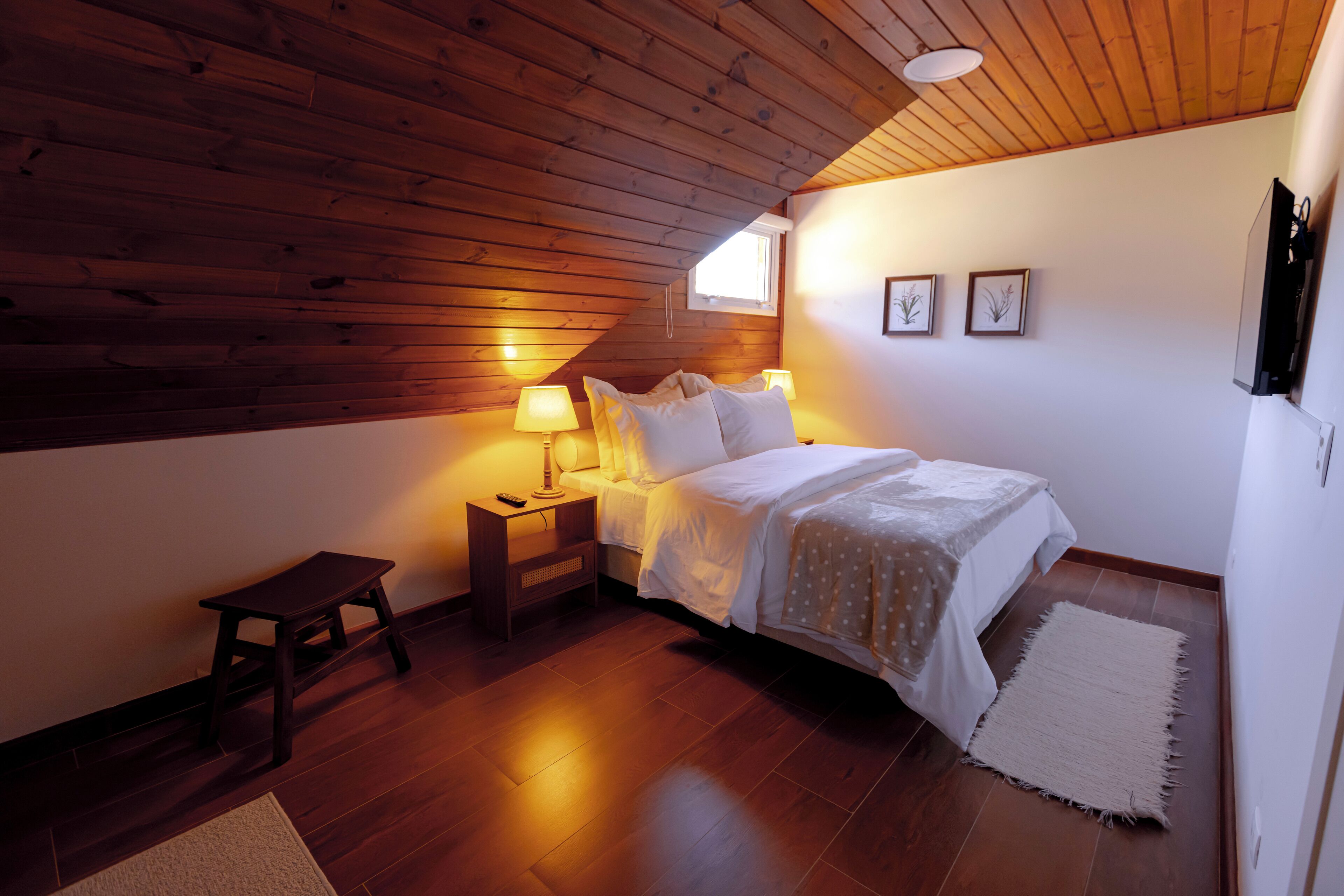 A bedroom with a wooden ceiling, a bed with white bedding, two bedside lamps, a small wooden stool, and a rug on the floor.