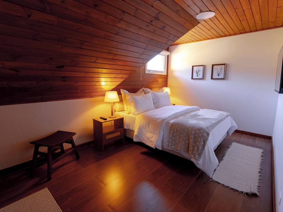 A bedroom with a wooden ceiling, a bed with white bedding, two bedside lamps, a small wooden stool, and a rug on the floor.