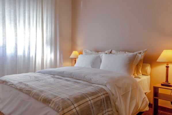 A neatly made bed with a plaid blanket, two bedside lamps, and a window with sheer curtains. - Zur Suítes (Campos do Jordão)