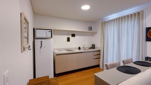 A compact kitchen with a refrigerator, microwave, and sink, a dining table with chairs, and a window with curtains. - Zur Suítes (Campos do Jordão)