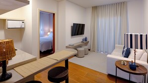 A modern hotel room with a sofa, a small table, a TV, a microwave, and a window with curtains. - Zur Suítes (Campos do Jordão)
