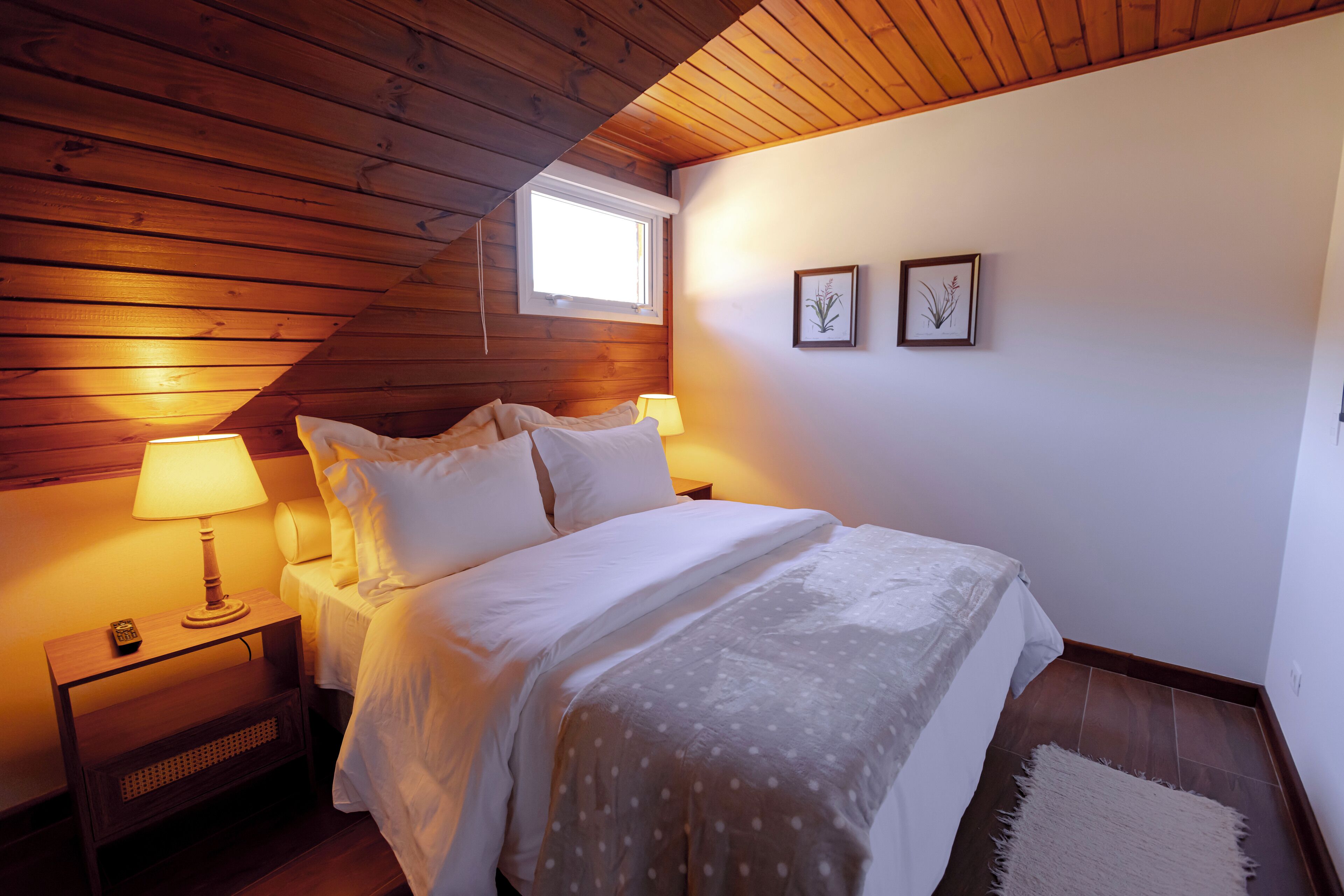 A bedroom with a wooden ceiling, a single bed with white bedding, two bedside lamps, and a small window.