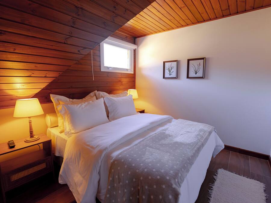 A bedroom with a wooden ceiling, a single bed with white bedding, two bedside lamps, and a small window.